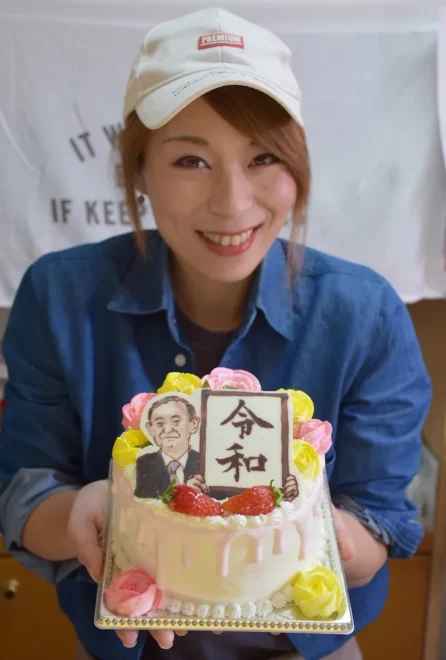 「​令​和​」​の​元​号​発​表​の​様​子​を​再​現​し​た​ホ​ー​ル​ケ​ー​キ​を​持​つ​池​田​さ​ん​。​官​房​長​官​の​表​情​は​、​微​妙​な​色​彩​で​描​き​上​げ​た​