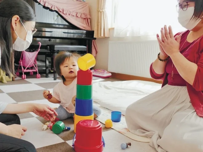 《１》「札幌市内の自宅で1歳の次女とカップ遊びをするネウボラ代表の坂本さん（左）。理事長の五嶋さん（右）も一緒に楽しみました」
